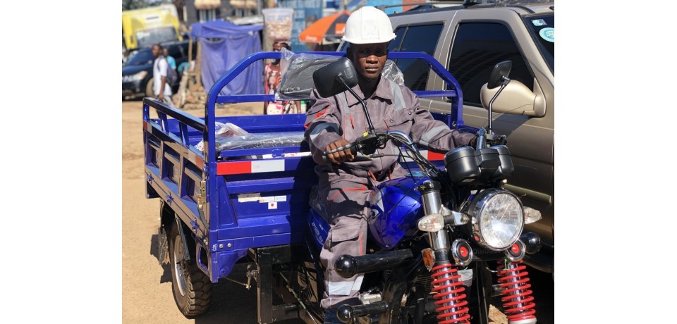 A participant in the Waste Management Micro-Enterprise Program, wearing a hardhat and uniform, drives a small vehicle down a crowded street