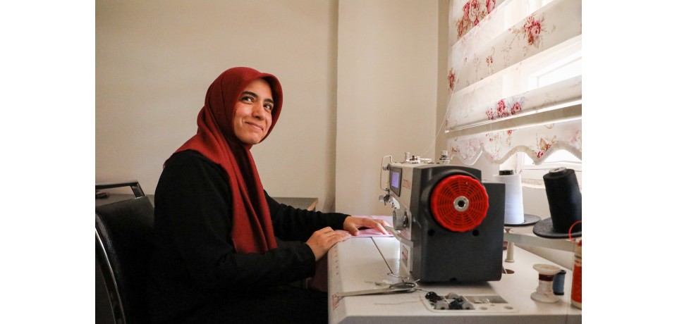 An image of a woman and small business owner sitting in front of a sewing machine and smiling at the camera
