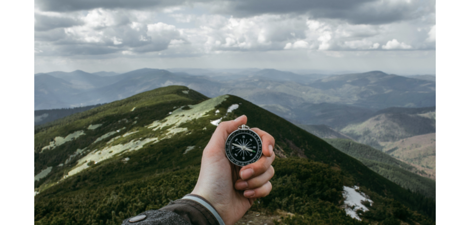 An image of someone's hand holding a compass, overlooking a mountainous landscape with clouds overhead