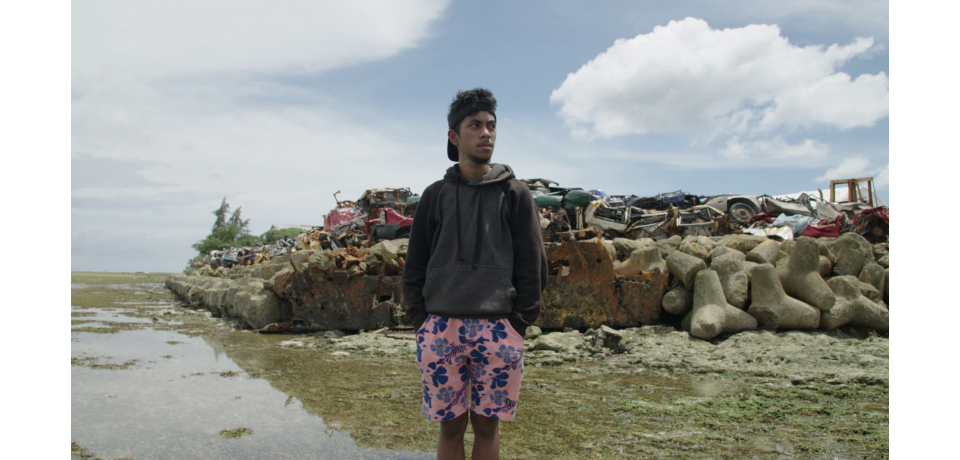 Teenage boy stands infront of a constructed sea wall made of old cars and blocks of cement