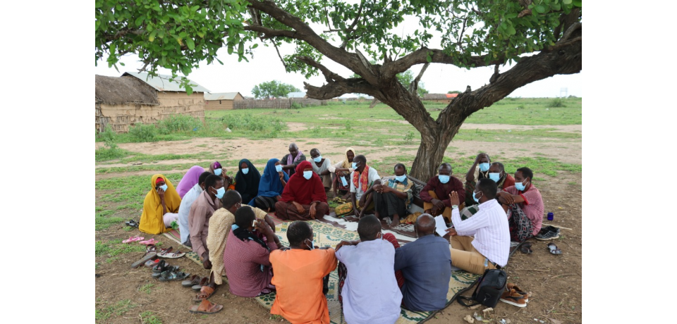 A group of people from a community, including men and women, sit under a tree and discuss