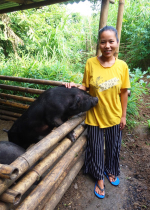 A woman standing next to a pigsty, petting a pig and smiling to the camera