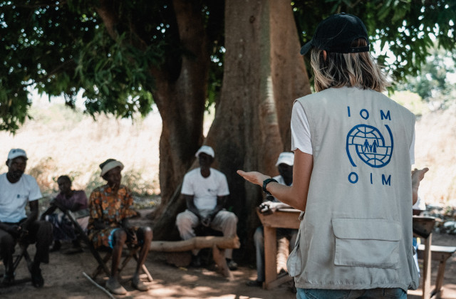 An IOM staff member discusses with the community sitting in chairs around a tree
