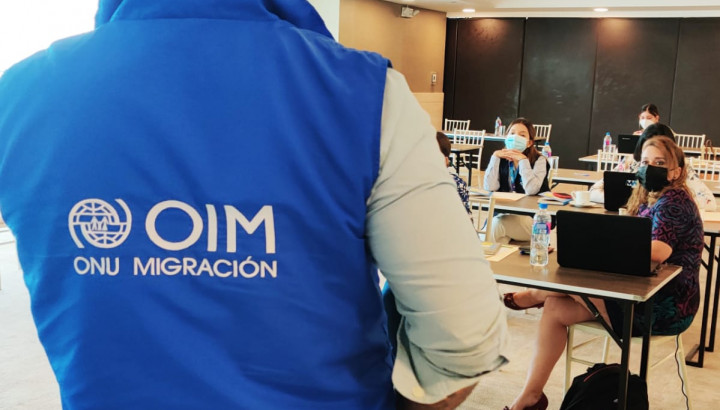 A man in blue vest with IOM logo speaking to people who are sitting in the meeting room